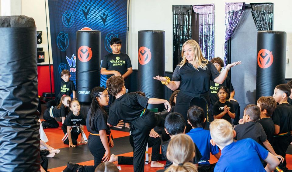 A regional director leads a class of kids on the mat in front of the Victory backdrop