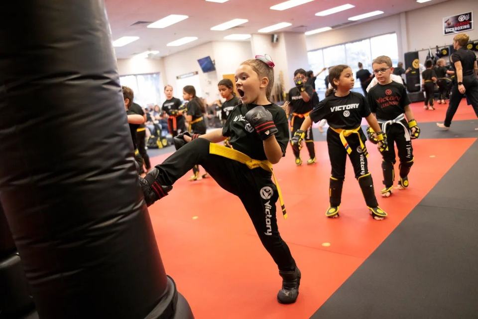 A girl in a black Victory uniform and yellow belt throws a high kick in sparring gloves and shouts with effort, her class lined up behind her on a red mat