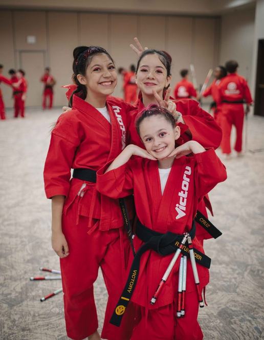 Three girls in red Victory uniforms and black belts pose close together, laughing, one making bunny ears; a tournament hall behind