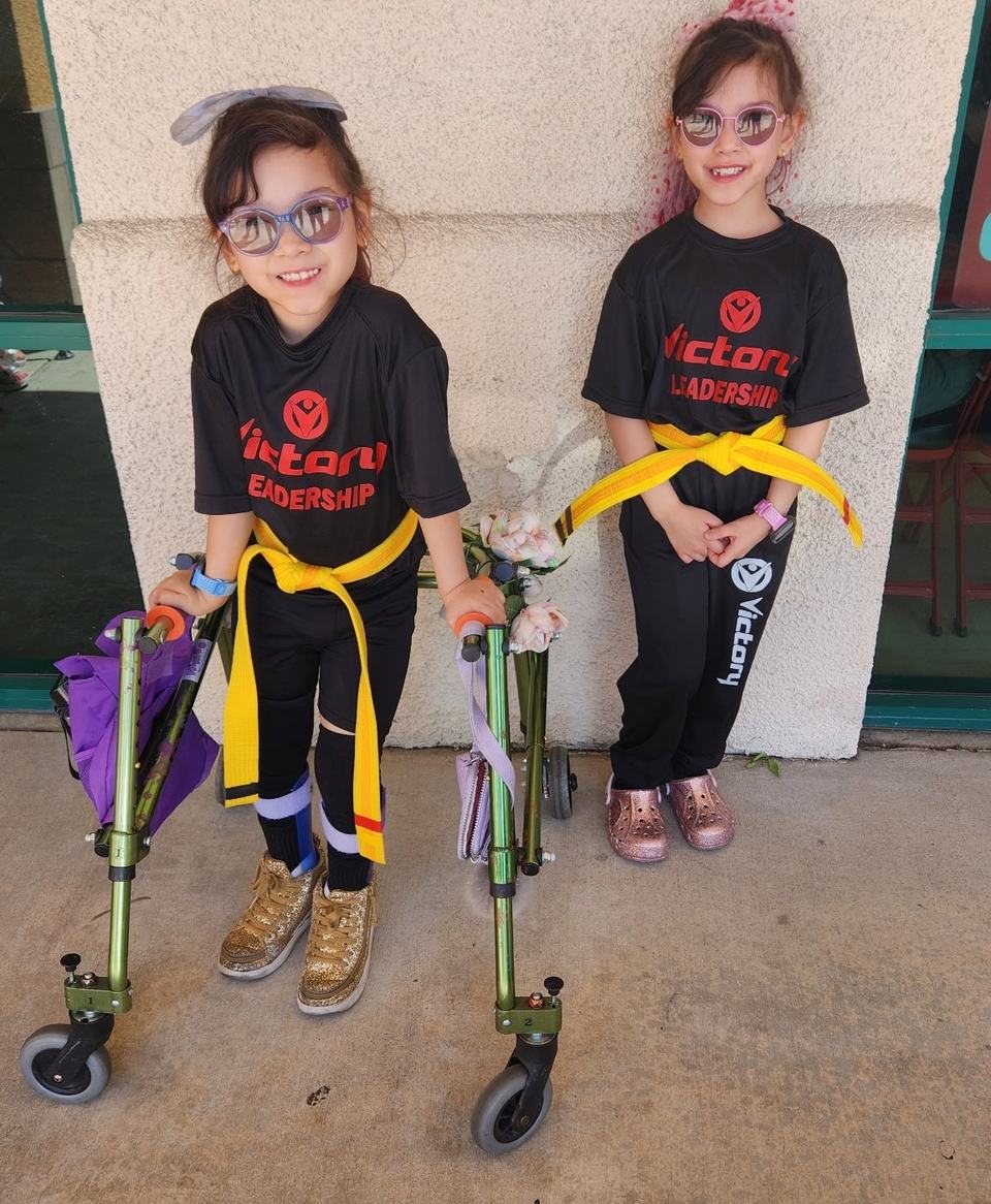 Twin sisters in Victory Leadership shirts and yellow belts smile against a studio wall; one stands in her green walker