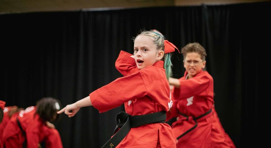 A girl black belt in a red uniform points and strikes a sharp pose, a classmate in red training right behind her