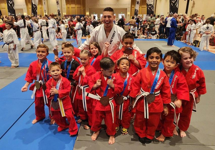 A beaming instructor stands among a team of little students in red Victory uniforms, all wearing medals